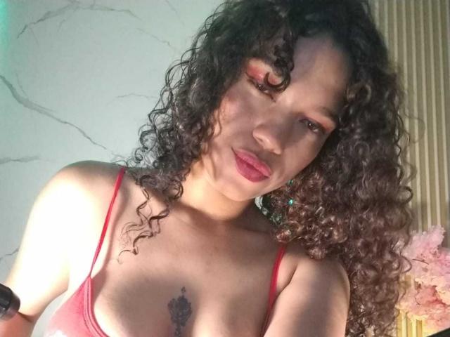 KellyAmbaris with curly brown hair, red lipstick, and red tank top, showing her tits and a tattoo under her boobs. She's looking down with a slight smile.