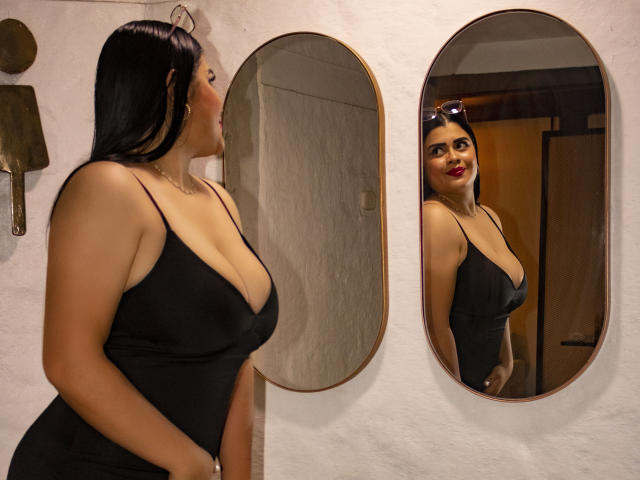 EmilyBoobsss with dark hair, red lipstick, and large tits in a black dress stands in front of an oval mirror, reflecting her smiling face and cleavage.