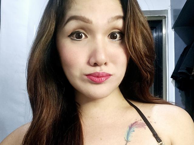 LovelyPinay69 with brown hair, pink lipstick, and a feather tattoo on her right shoulder, looking straight at the camera with a neutral expression.