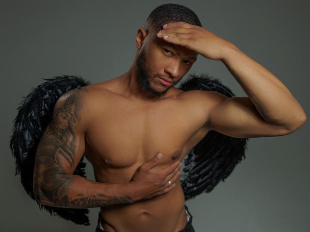 JadenSmithy, shirtless, black feathered wings, tattooed arm, dark skin, short haircut, beard, hand covering chest, looking down, gray background, wearing black pants, serious expression, ring on finger.