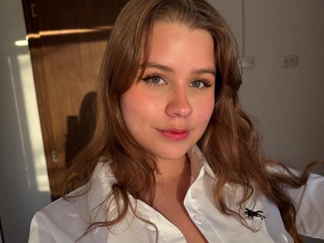 SwanKyivy heeft lang bruin haar, groene ogen en roze lippen. Ze draagt een witte blouse met een zwart logo. Haar borsten zijn licht zichtbaar, en ze glimlacht..
