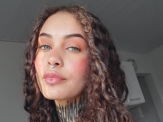 CasalMuitoQuente has curly brown hair, green eyes, a nose ring, and a lip piercing. Their face is slightly flushed with pink cheeks, wearing a black turtleneck.