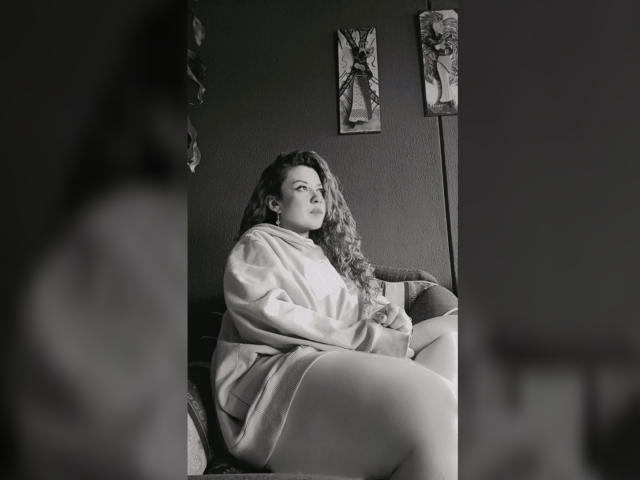 Black-and-white photo of RosseHot6. RosseHot6, a curvy woman with long, wavy hair, sits on a dark couch. She wears a loose, light-colored robe that covers her upper body and thighs. Her expression is neutral, with eyes slightly upward. Two abstract paintings hang on a dark wall behind her. The image uses soft, diffused lighting. RosseHot6's body is positioned slightly to the right, with her knees up and her hands resting on them. The composition is framed by blurred elements in the foreground.