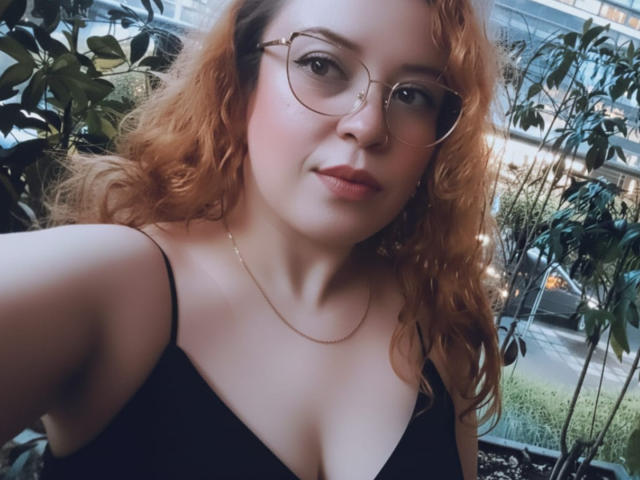 RosseHot69 ha i capelli rossi ricci, degli occhiali e un vestito nero con un décolleté basso che mostra il seno. Indossa una catena d'oro e guarda la fotocamera con un leggero sorriso..