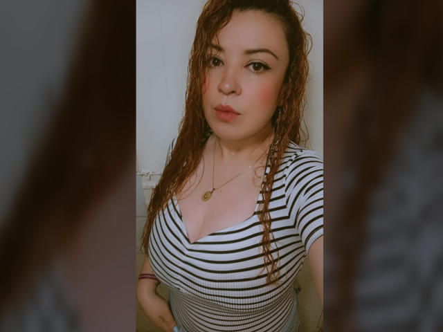 RosseHot69 con capelli castano ricci, indossa una camicia a righe, mostra un seno prosperoso, labbra piene e una collana d'oro, ed è in piedi di fronte a uno specchio sfocato..
