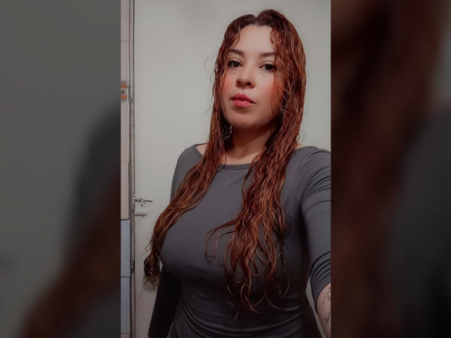 RosseHot69 mit nassen, lockigen braunen Haaren, einem grauen Langarmshirt und vollen Lippen, steht in einem Raum und spiegelt sich in einem Spiegel wider, wobei ihre Brüste und ihr Hintern zu sehen sind..