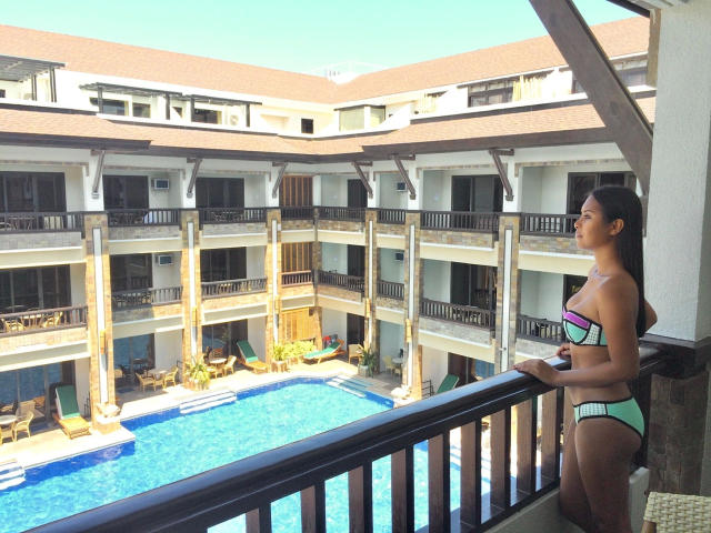 TsLexie-hot standing on a balcony, wearing a green and pink bikini, looking at a blue pool, with her ass and tits visible, under a clear sky.