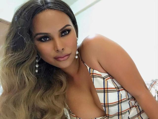 TsLexie-hot with long, wavy brown hair, big blue eyes, and a plaid dress showing her tits. She's wearing pearl earrings and giving a sultry look.