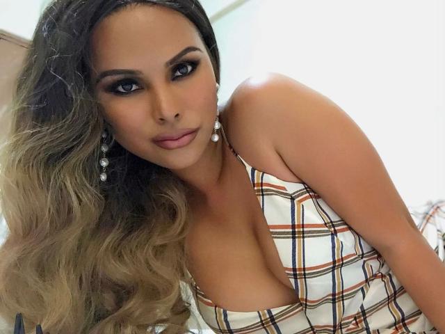 TsLexie-hot with big tits in a plaid top, looking at the camera with dark makeup and long, wavy blonde hair, wearing dangling earrings, in a soft light.