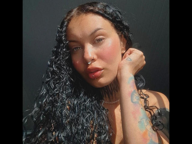 CasalMuitoQuente has curly black hair, a nose ring, and lip ring, with a tattoo on their right arm and a necklace, against a dark background.