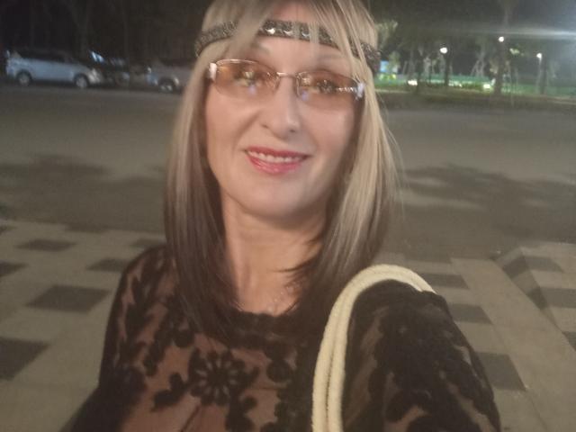 ElizaWendy with glasses, wearing a black lace top, smiling, headband, blonde hair, white bag, night, street, car in background. Her tits are slightly visible through the lace.