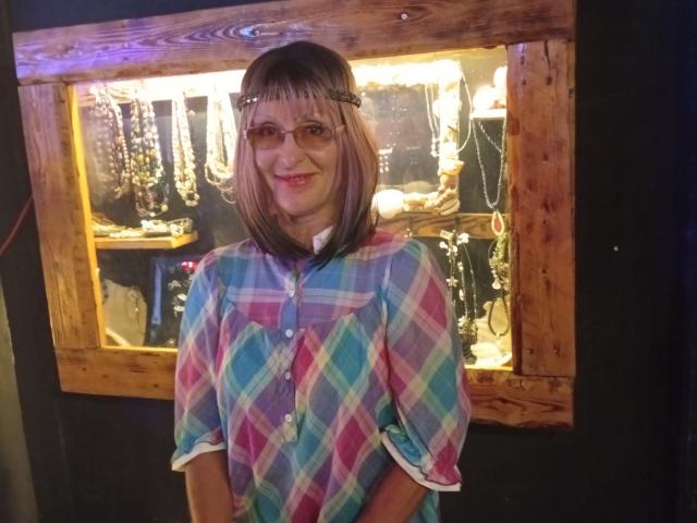 ElizaWendy with pink-tinted bob, glasses, smiling, wearing a colorful plaid shirt, standing in front of a wooden-framed jewelry display.