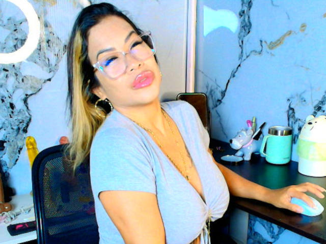 AlixLow with pouty lips, wearing glasses, gray top, and gold necklace, has her hand on a mouse, blonde hair, marble walls, and a ring light in the background.