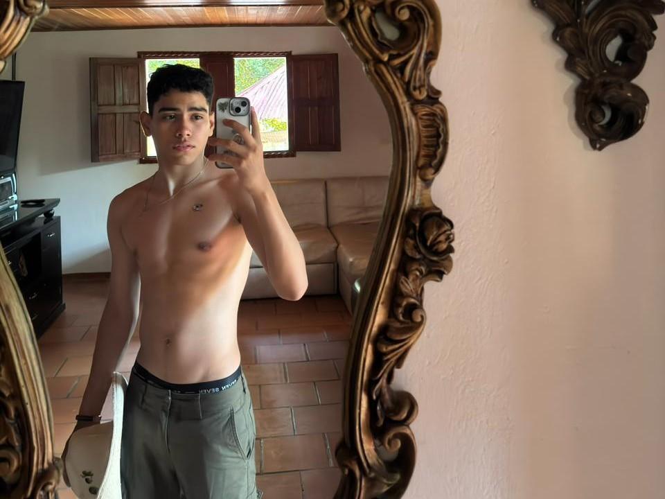 Shirtless Yamilk69 with a slim, toned body, dark short hair, and olive skin stands in front of an ornate mirror, holding a phone, wearing green pants, and black underwear.