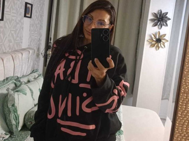 CelestePoot con gafas, cabello castaño, sudadera negra de gran tamaño con letras rosas, sosteniendo un teléfono, selfie frente al espejo, de pie en un dormitorio, almohadas verdes, cama blanca, cortinas grises..