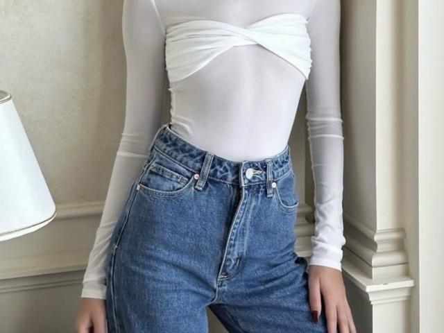 GlariyaHot has white, wrap-style, long-sleeve top, showing her tits, and wears high-waisted, blue, button-up jeans with a visible button. Her hands rest on her thighs.