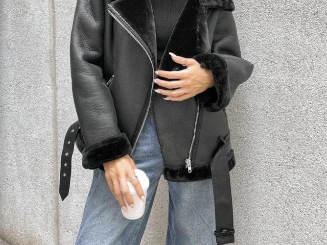 GlariyaHot wearing a black leather jacket with fur cuffs, blue jeans, and holding a white phone. Black belt hangs from jacket. Hands visible, nails painted white.