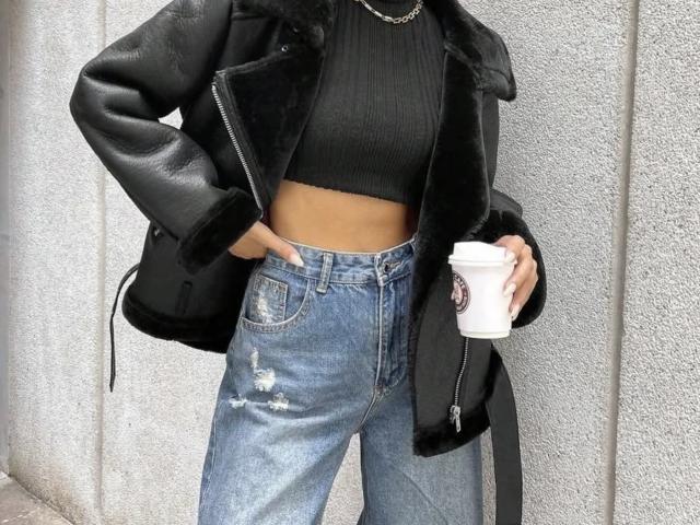 GlariyaHot with black leather jacket, black ribbed crop top, blue distressed jeans, holding white coffee cup, black backpack, wearing chain necklace, standing against grey textured wall.