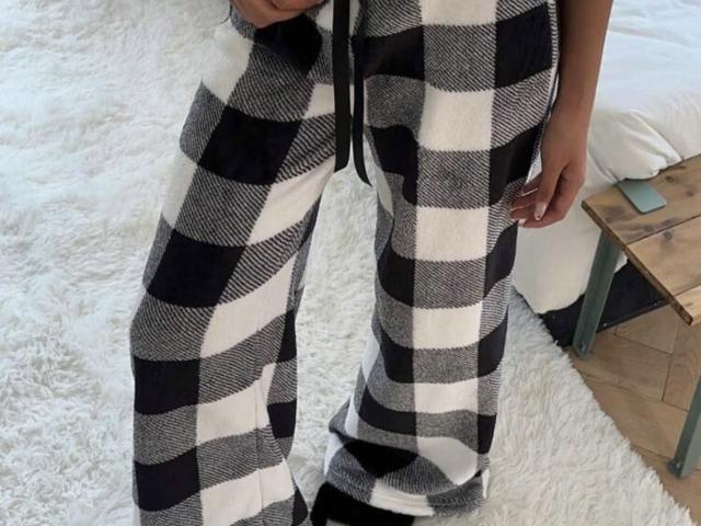 GlariyaHot wearing black and white checkered pants, standing on a white fluffy rug, with a wooden table and pillow in the background. Pants are flared, black belt visible.