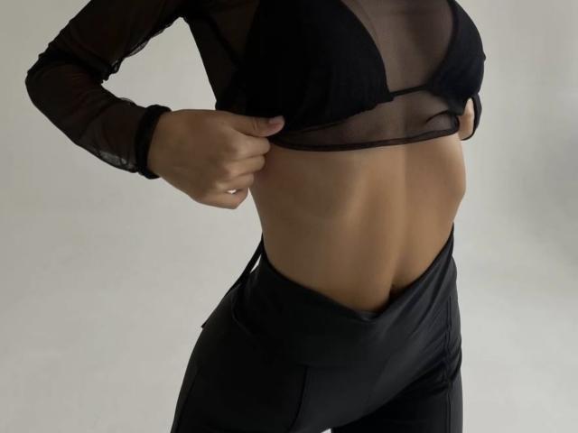 GlariyaHot's hands pull up a sheer black top, revealing small, black-straped boobs and a toned, flat, tan midriff with black, high-waisted pants.