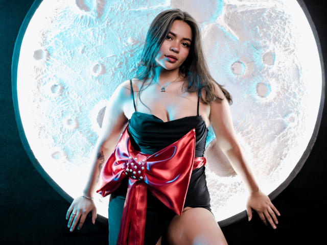 KimDevinne with medium tits, wearing a black dress and red satin bow, standing against a glowing moon-like circular light, hands on her thighs, blue highlights in her hair.