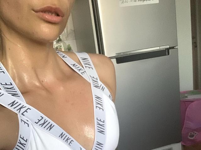 BellaRayyy's upper body in a selfie. She has shiny, sweaty skin and wears a white Nike sports bra with black "NIKE" text on the straps. Her lips are slightly parted, and her face is partially visible. The background includes a gray refrigerator and a pink backpack. The image focuses on her chest and lips. The composition is close-up. Her skin is light, and she has a subtle, natural look. The lighting is even.