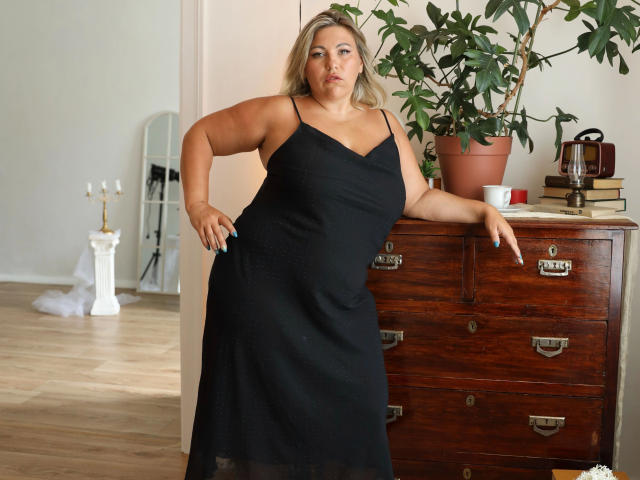 AlysandraSex-hot stands in a black dress, arms slightly raised, hand on wooden dresser, showing her ass and large tits, with a potted plant and candles behind her.