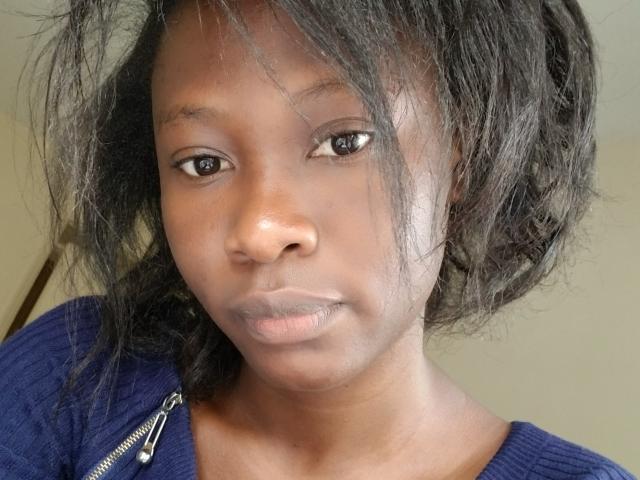 PrettyM's face, dark brown skin, black wavy hair, small nose, serious expression, wearing a blue zip-up shirt, visible zipper, slightly parted lips, looking at the camera.