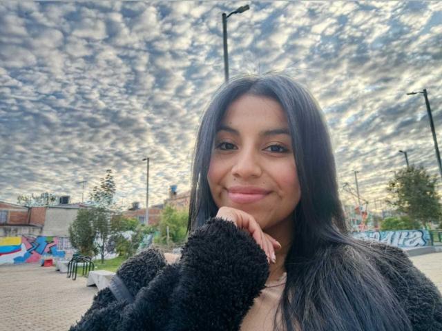 EmmaSollins has long black hair, a cute smile, and is wearing a fluffy black sweater, with a cloudy sky behind her. Her hand is near her face, showing off nails.