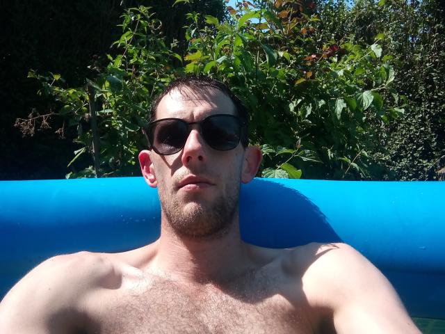 RainySexy, shirtless, with dark sunglasses, has a hairy chest, short brown hair, and a serious face, lounging in a blue inflatable pool under sunny green leaves.
