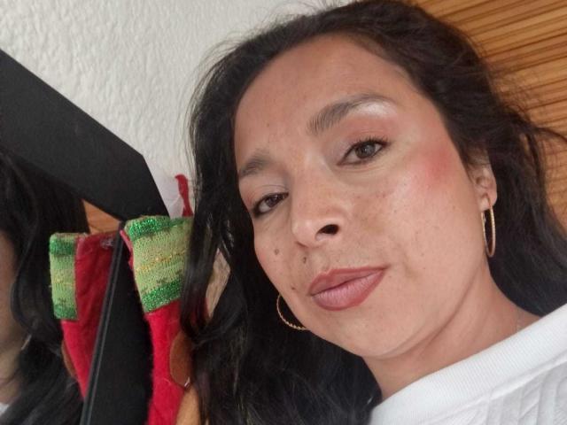 StangelarRivera with puffy red lips, brown eyes, and black wavy hair, wearing gold hoop earrings, holding a red and green cloth, smiling slightly. Her face has small moles.