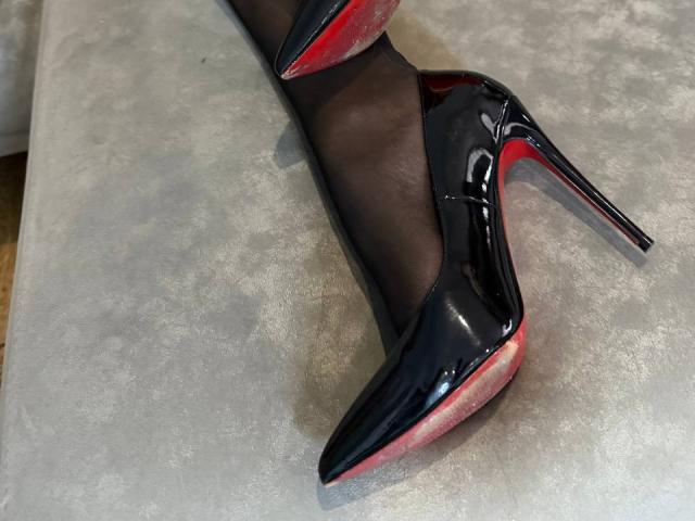Close-up of Niferttiti's foot in a shiny black patent leather stiletto heel with red soles. The heel is pointed up, showing its sleek, glossy texture. Niferttiti is wearing sheer black pantyhose. The background is a gray, slightly textured surface. The image highlights the contrast between the black heel and red soles, with the glossy patent leather reflecting light. The composition focuses on the elegance and sharpness of the stiletto.