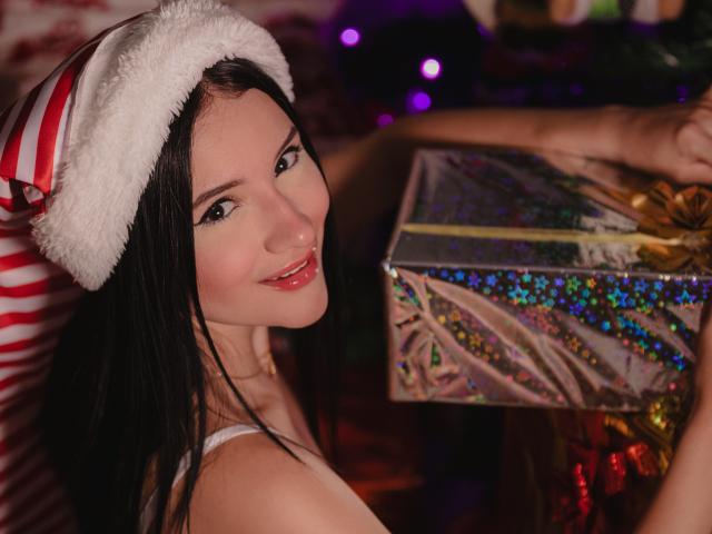 LizzJoy with long black hair, wearing a Santa hat, smiling at the camera, holding a glittery, colorful gift box in front of her, showing her bare shoulders.