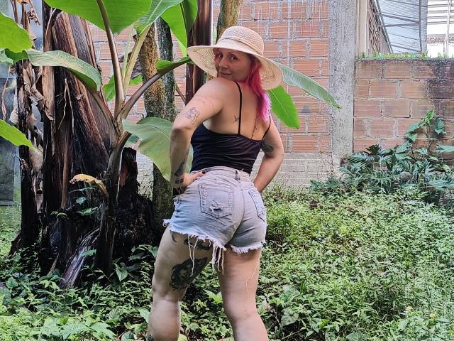 GiselleDunn with pink hair, straw hat, black tank top, and frayed denim shorts, showing her ass and tattoo on her right thigh, standing by a green plant.