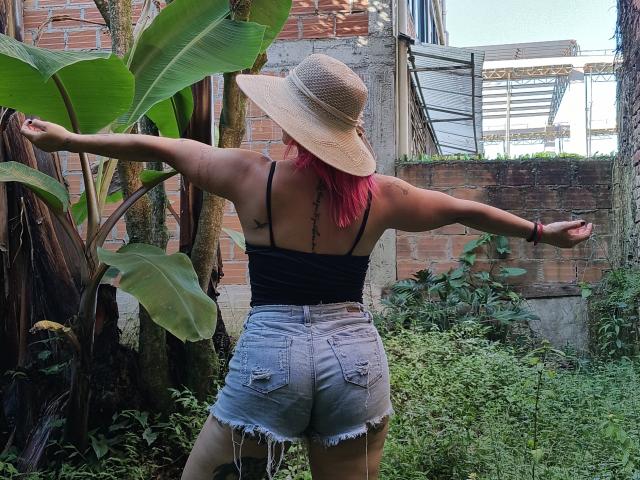 GiselleDunn stands with arms out, back to camera, wearing a sun hat, black top, and frayed denim shorts, pink hair, showing her ass, in a green, leafy yard.