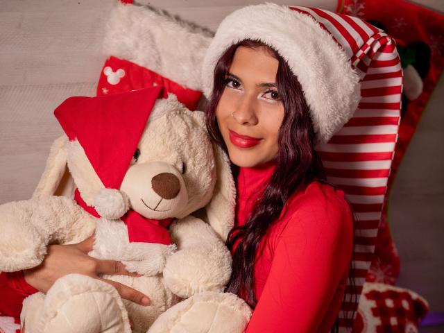 DanikaSerov with red lipstick, wearing a Santa hat, hugging a white stuffed bunny with a red bow, in a red top, smiling at the camera.