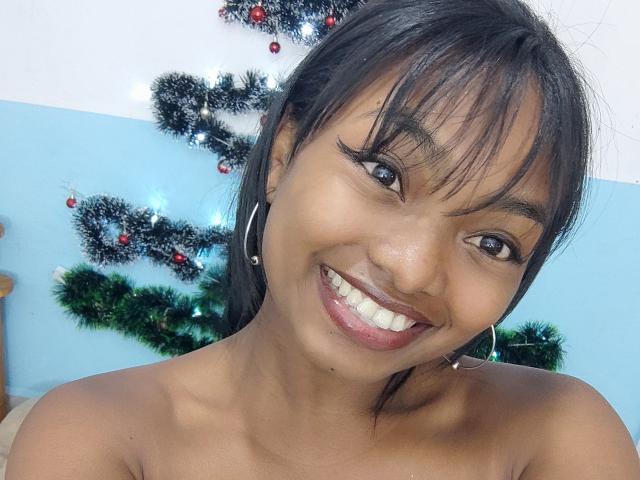 Urshula, smiling with dark brown eyes, black hair, and hoop earrings, showing her teeth and pink lipstick, topless with her small, dark brown tits visible.