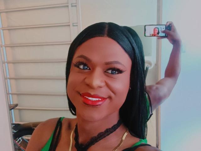 VenusMiller with dark skin, red lipstick, black hair, green top, holding phone in mirror, smiling, showing her tits, black lace choker, gold necklace, selfie.