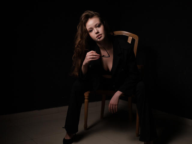 BeatriceKeat sits in a wooden chair against a dark background. She has long, wavy brown hair and a serious expression. She wears a black jacket, black pants, and black flats. Her right hand holds black-framed glasses near her chest. The lighting is low, creating shadows on her face and highlighting her left cheek. Her left arm rests on the chair's armrest. The floor is light gray, contrasting with the dark background and her clothing. The image uses chiaroscuro to emphasize her facial features and the texture of her hair.