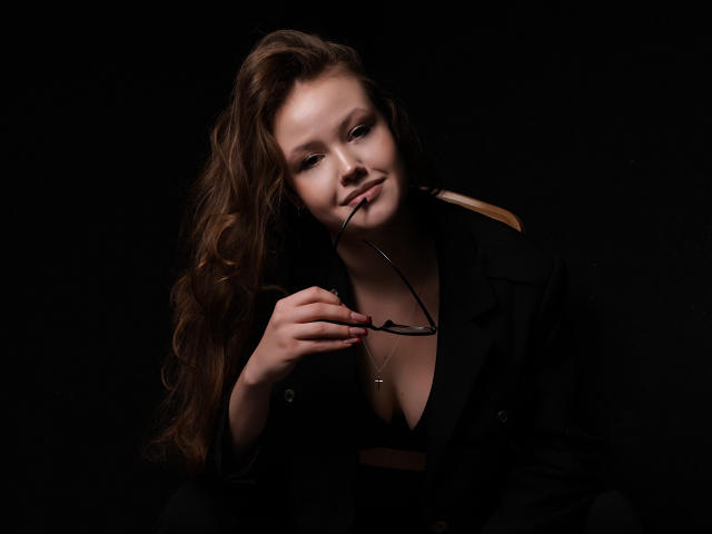 BeatriceKeat with long, wavy brown hair sits against a dark background. She holds black-rimmed glasses in her right hand, lightly biting the left arm. She wears a black jacket, exposing a low neckline with a gold cross necklace. Her skin is fair, and she has a subtle, confident smile. The lighting is low and dramatic, highlighting her face and hair while keeping the rest in shadow. The image has a realistic, high-contrast style.