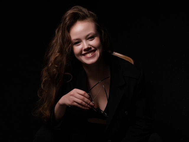 BeatriceKeat with long brown hair, smiling, wearing a black jacket, holding glasses in her right hand, dark background, soft lighting, showing her tits under the jacket.