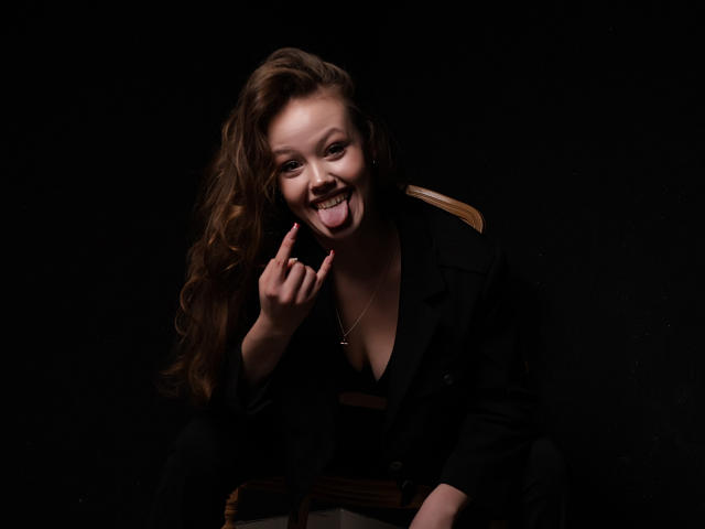 BeatriceKeat with long brown hair, black jacket, and necklace, sticking out tongue, winking, and pointing at camera, dark background, looking sexy and playful.