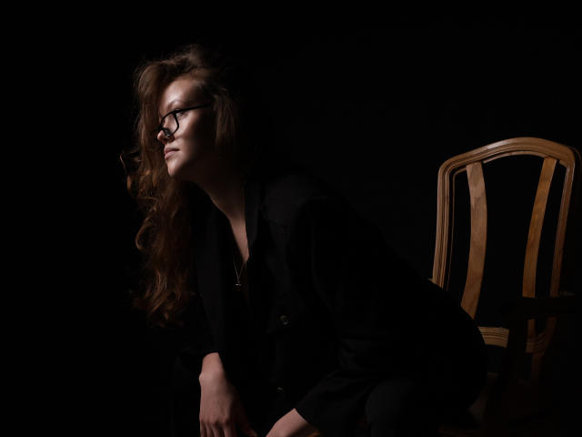 BeatriceKeat with wavy brown hair, glasses, black shirt, and black pants, leaning on a wooden chair, dark background, side profile, looking to the left.