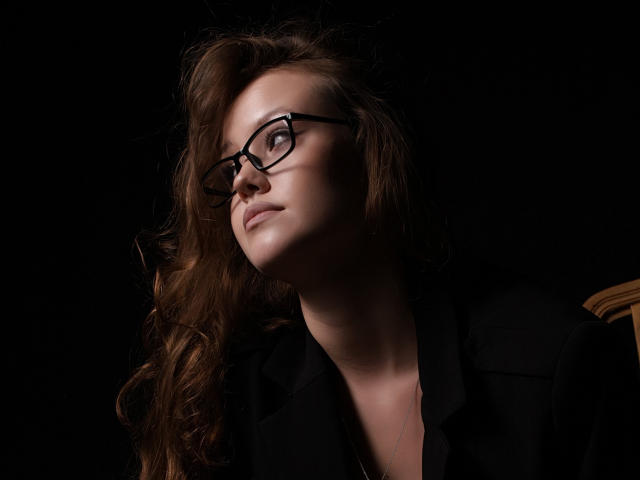 BeatriceKeat with wavy brown hair, black glasses, and black shirt, looking up with a slight smile, dark background, soft lighting, face in shadow, upper body shot.
