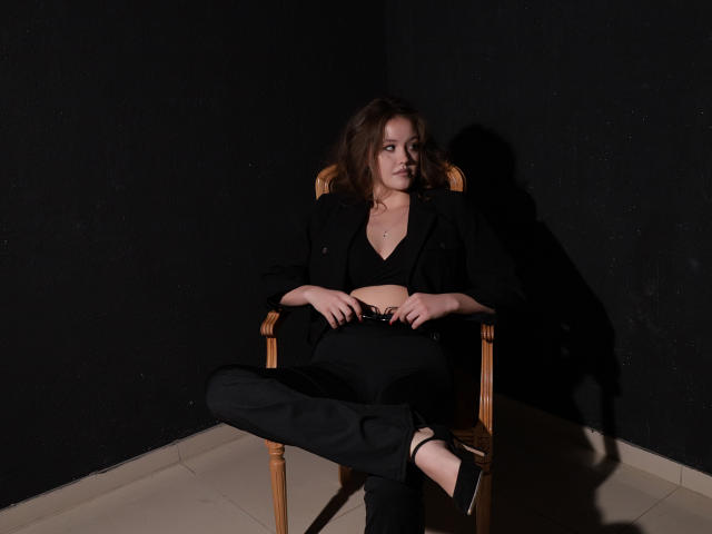 BeatriceKeat is seated in a wooden chair with dark black walls and a tiled floor. She has medium-length brown hair, a fair skin tone, and a slender build. She's wearing a black crop top, black pants, and black strappy heels. Her hands rest on her lap, and her legs are crossed. The lighting is low, casting a shadow on the wall behind her. The composition is simple and centered, emphasizing her relaxed posture. The textures include the wooden chair, her clothing, and the smooth tiled floor. The overall style is minimalistic and focused.
