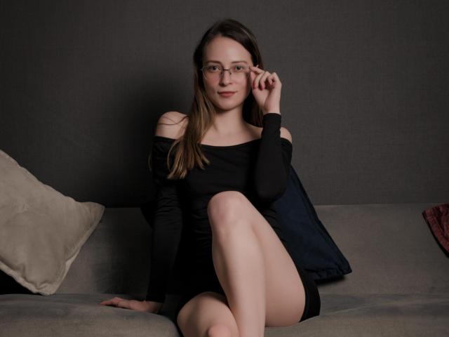 AmbeerWood with glasses, black off-shoulder dress, and bare shoulders, sitting on gray sofa, legs crossed, right hand near face, light skin, brown hair, subtle smile.