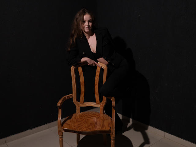 BeatriceKeat with wavy brown hair, black outfit, and slight smile, sitting on a wooden chair, hands on top, in dim lighting, shadows on dark wall, tile floor.