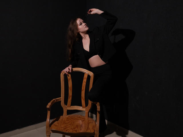 BeatriceKeat with wavy brown hair, black outfit, and midriff, leaning on a wooden chair, right arm raised, shadow on dark wall, white tiled floor.
