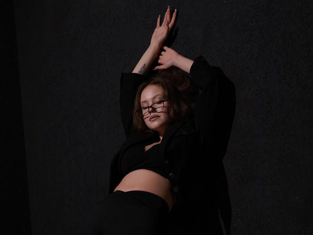 BeatriceKeat with glasses, arms above head, wearing black shirt and black pants, has a tattoo on her right wrist, mid-shot, looking slightly down, dark background.