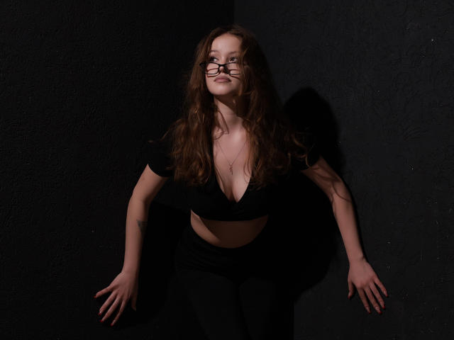 BeatriceKeat with long brown hair, wearing glasses, black crop top, and black pants, leaning against dark wall, hands down, small tits, subtle cleavage, tattoo on left arm.